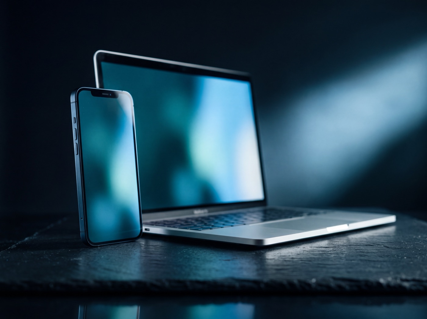A smartphone standing beside an open laptop on dark slate, both screens lit
